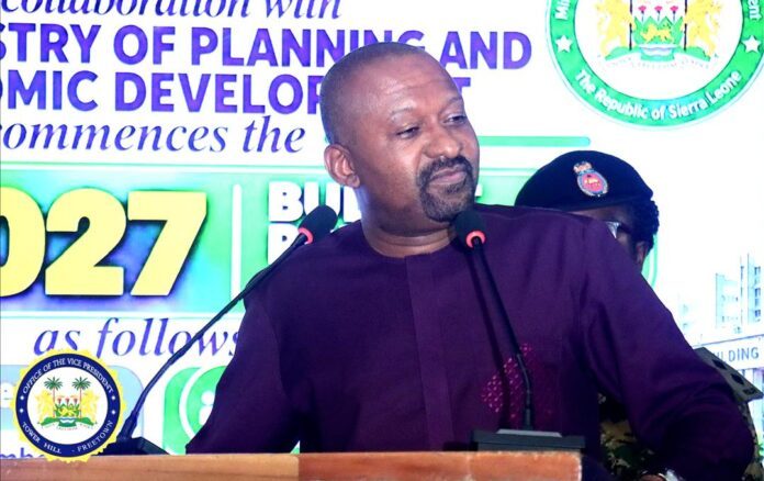 Man in a purple traditional outfit delivering a speech at a podium with two microphones, against a backdrop of banners and emblems.