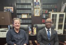 British High Commissioner Applauds Judiciary’s Ongoing Reforms Two professionals sit in leather chairs in a dark wood library, facing the camera; woman in navy dress, man in a pinstripe suit with a blue tie.