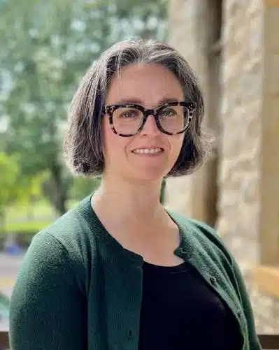 Smiling woman with short gray hair and glasses standing outdoors beside a stone wall.