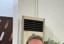 Hotel Association President John Shallop Appointed to National Tourist Board Smiling man in a blue suit with a headset sits at a desk with a laptop, mug, and a potted plant behind him.