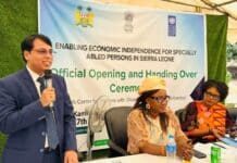 India-Funded Skills Training Centre Opens in Kambia to Empower Persons with Disabilities Man in a blue suit speaks into a microphone at a panel, with a banner about enabling economic independence for specially abled persons in Sierra Leone behind him.