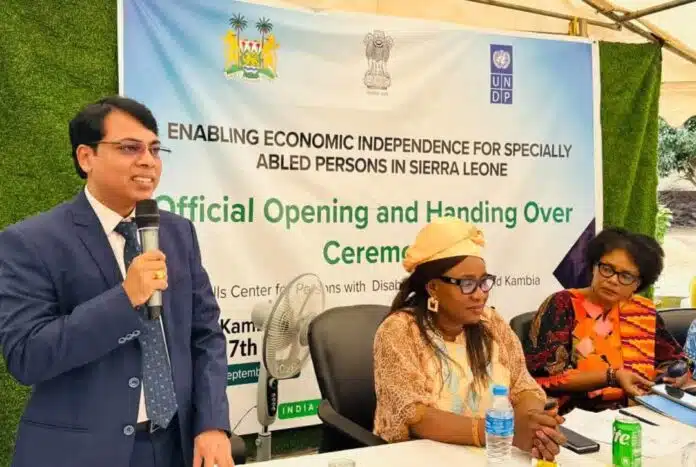 Man in a blue suit speaks into a microphone at a panel, with a banner about enabling economic independence for specially abled persons in Sierra Leone behind him.