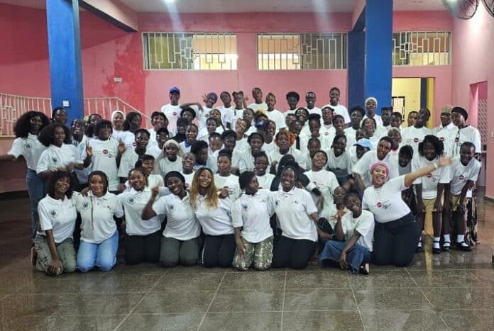 Large diverse group of youths in white shirts posing for a group photo in a pink-walled hall with blue columns and barred windows in the background.