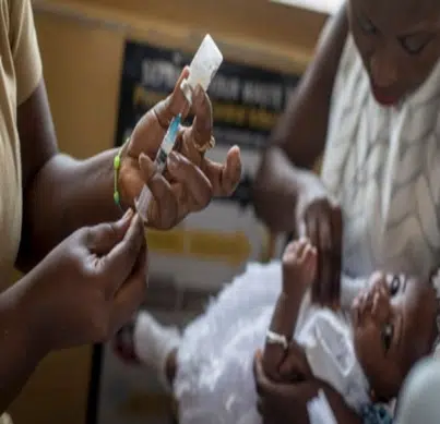 Healthcare workers administer a vaccine to a baby during a clinic visit, with one person holding the baby and another preparing a syringe.