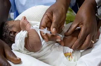 Newborn baby wrapped in a white blanket, held by caregivers as a clinician prepares to give a neonatal injection.