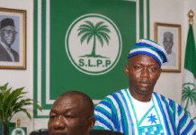 As He Sends Strong Message to President Bio… Hon. Alhaji Musa Tarawally Fires Back at Batilo Songa, Insists He’s a Paid-Up Member Two men in a formal office: one seated at a wooden desk in a light blue traditional outfit with a gold watch, the other standing beside him in blue‑green striped attire, frente a a green SLPP emblem backdrop.