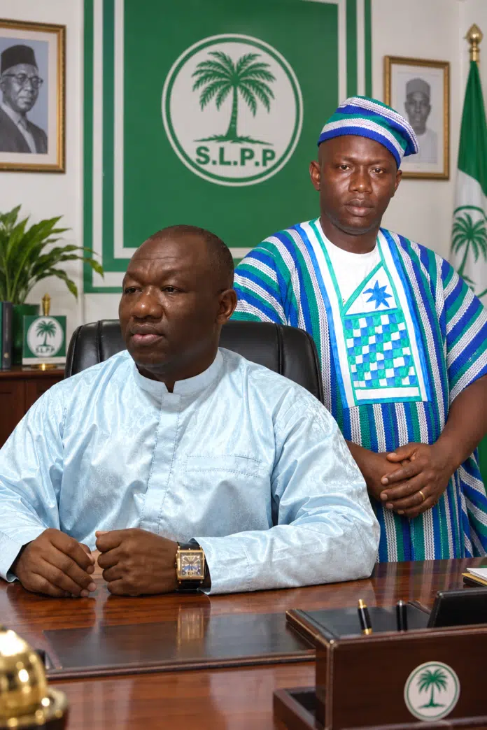 Two men in a formal office: one seated at a wooden desk in a light blue traditional outfit with a gold watch, the other standing beside him in blue‑green striped attire, frente a a green SLPP emblem backdrop.