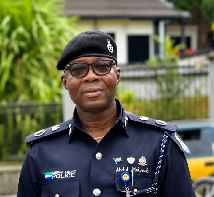 Portrait of a Sierra Leone police officer in navy uniform and beret, standing outdoors with greenery in the background.