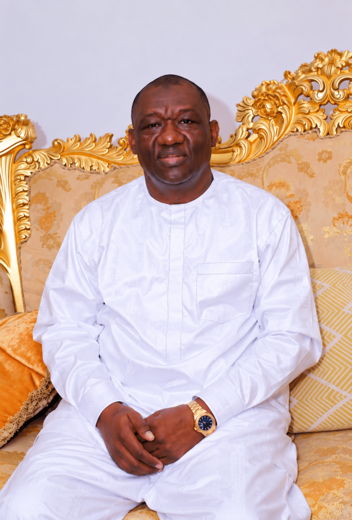 Portrait of a man in a white traditional outfit seated on an ornate gold couch with patterned cushions, hands folded in his lap.