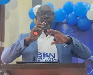 Man in a blue blazer speaks at a podium, holding an orange microphone, with blue balloons behind him at the event evening backdrop.