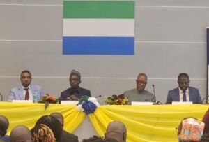 Panel of four men seated at a long table draped in yellow, with flower arrangements, facing an audience; Sierra Leone flag on the wall behind them.
