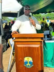 Man in a light traditional shirt speaks into a microphone at a wooden podium under a green canopy, with an audience seated behind him.
