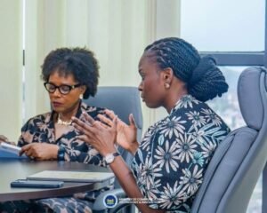 Two professional women in a meeting: one wearing glasses taking notes, the other speaking with hand gestures; a government ministry logo visible on the table.