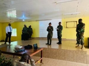 Soldiers in uniform giving a briefing in a yellow-walled room with a projector screen at the front.