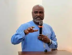 Man in a blue traditional outfit speaking into a handheld microphone at a wooden podium, gesturing with his left hand as he talks to an audience.