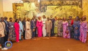 Group of men and women in colorful traditional outfits posing together in a hall with a gold-toned mural backdrop, logo visible in the corner.