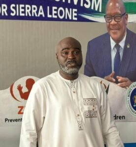 Man in a light beige traditional outfit stands in front of a banner about Sierra Leone, with a suited official image in the background.