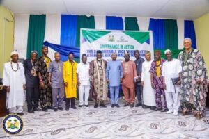 Group of men in colorful traditional outfits standing on a decorated stage in front of a banner that reads 'Governance in Action' during a formal event, with green, white, and blue drapes and an official seal visible in the bottom-left.