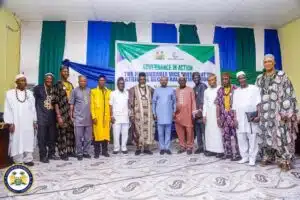 Group of men in colorful traditional outfits standing on a decorated stage in front of a banner that reads 'Governance in Action' during a formal event, with green, white, and blue drapes and an official seal visible in the bottom-left.
