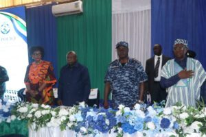 Dignitaries stand behind a long table with a blue-and-white flower garland on a formal stage.