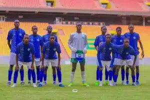 Soccer team posing for a group photo on the field, goalkeeper in gray jersey center holding a banner, players in blue jerseys and white shorts.