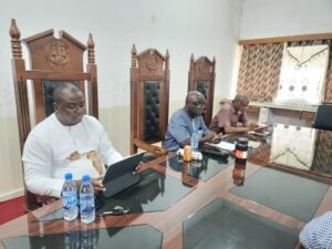 Three men sit at a long conference table with ornate wooden chairs, reviewing documents on tablets and notebooks in a meeting room.
