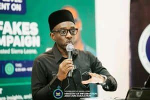 Man in a black shirt and glasses speaks into a microphone at a campaign event, with a green banner reading 'FAKES CAMPAIGN' in the background.