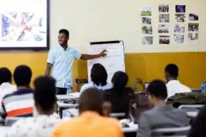 Male instructor in a light blue shirt points to a notes-filled flip chart while students sit at desks in a bright classroom-room setting.