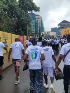 Crowd of people in white T-shirts walking down a wet city street during a peace walk, yellow barriers on the left and tall buildings ahead.