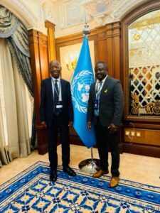 Two men in dark suits stand beside a United Nations flag in an ornate room, posing for a formal photo (both wearing lanyards).