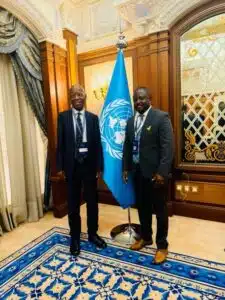 Two men in dark suits stand beside a United Nations flag in an ornate room, posing for a formal photo (both wearing lanyards).