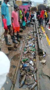 People stand along a street beside a clogged gutter filled with plastic bottles and trash, observing the mess ahead.