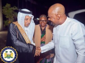 Two men shake hands at night; one wears traditional Arab dress, the other a light tunic, with a smiling woman in the background and an official-looking seal in the corner.