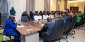Formal meeting in a conference room with a long table and many attendees; a man in a blue suit sits at the head of the table