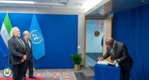 Two men in suits stand beside a signing table during a formal ceremony; UN flag and another flag behind them.