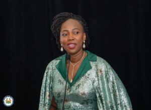 Woman in a green sequined dress speaks into a microphone on a dark stage, wearing pearl earrings and a necklace.