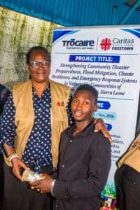 Woman in a beige vest and glasses hands a small wrapped gift to a young man in a black shirt, beside a banner for Trocaire/Caritas Sierra Leone project.
