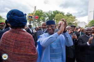 Smiling man in light blue traditional suit and cap claps among a crowd at an outdoor ceremony.