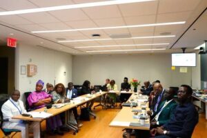 Diverse group of professionals seated around U-shaped conference tables in a bright meeting room, with documents, laptops, and a wall-mounted screen in the background.