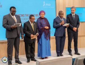 Group of five formally dressed individuals standing on a stage, each holding papers, at a blue-sponsored event backdrop.