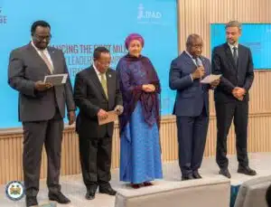 Group of five formally dressed individuals standing on a stage, each holding papers, at a blue-sponsored event backdrop.