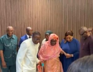 Group of well-dressed people circle an elderly woman in a pink traditional outfit, offering support at a formal event.