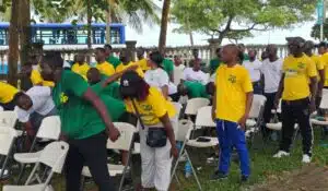 Group of people in yellow and green shirts gathering outdoors, some seated on white folding chairs as others stand or bend, with a blue bus and trees in the background.
