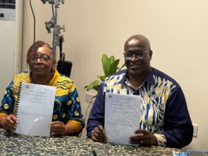 Two people sit at a patterned table indoors, each holding a document and looking at the camera; the woman wears a colorful patterned top, the man a blue-yellow shirt, both smiling slightly.