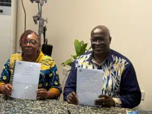 Two people sit at a patterned table indoors, each holding a document and looking at the camera; the woman wears a colorful patterned top, the man a blue-yellow shirt, both smiling slightly.
