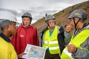 Four construction workers in hard hats and reflective vests study a site plan map outdoors near a rocky slope.
