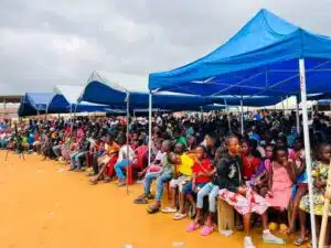 Large crowd seated under blue canopy tents at an outdoor event on dusty orange ground, with people of all ages watching something off-camera.