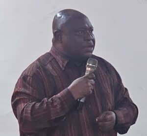 Man in a brown striped shirt holds a microphone and speaks on stage.
