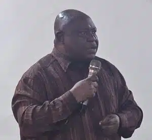 Man in a brown striped shirt holds a microphone and speaks on stage.