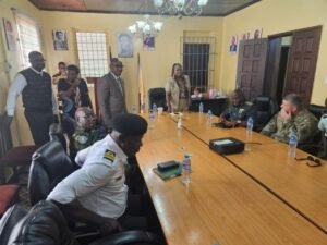 A conference room with a long wooden table, several uniformed officers seated and standing, and a woman speaking near the head of the table in a yellowish room. Water bottles and a projector sit on the table.